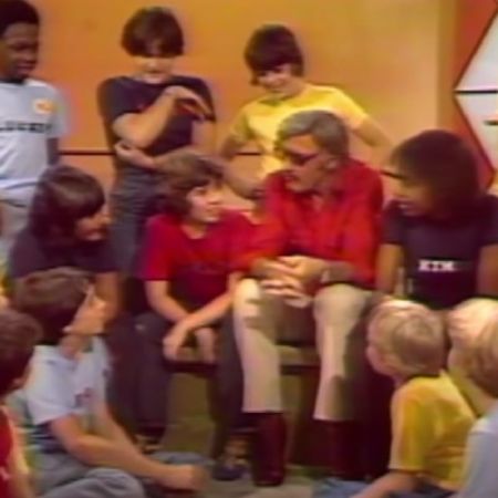 Stan Lee is surrounded by kids as he is talking to them.